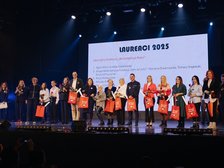Na zdjęciu widzimy grupę kilkunastu osób stojących obok siebie na scenie. W tle znajduje się duży biały ekran z czarnym napisem Laureaci 2025, poniżej wymienione imiona i nazwisk. Większość z nich trzyma w dłoniach pomarańczowe torby z białym napisem „Moje zakupy niosą pomoc” oraz czerwone róże. Dwie z nich dodatkowo trzymają puchar laureata. Uczestnicy są zwróceni przodem do widza i pozują do wspólnego zdjęcia.