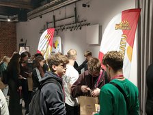 Na zdjęciu widzimy grupy młodzieży po trzy, cztery osoby, które ze sobą rozmawiają. Za nimi widzimy windery w kolorach białym, żółtym, czerwonym z żółtym napisem Ziemia Pomagająca. W tle widzimy białą ścianę z punktami świetlnymi. 