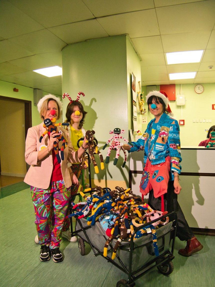 Na zdjęciu widać trzy dorosłe osoby przebrane w kolorowe stroje klownów. Stoją w szpitalnym korytarzu obok wózka pełnego pluszowych, materiałowych małpek.. Mają czerwone nosy i czapki, uśmiechają się i trzymają materiałowe maskotki w rękach