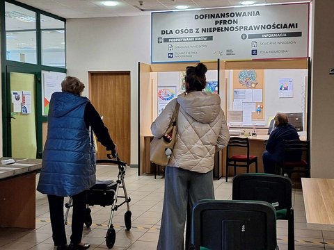 Sala obsługi MOPS w Łodzi. Kobieta wsparta o balkonik czeka w kolejce do okienka obsługi z napisem Dofinasowanie PFRON dla osób z niepełnosprawnościami