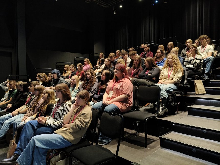 Zdjęcie przedstawta widownię w teatrze, która wypełniona jest młodzieżą. Na pierwszym planie mamy kilka pustych krzeseł i schody, którymi można wejść do wyższych rząedów.