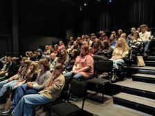 Zdjęcie przedstawta widownię w teatrze, która wypełniona jest młodzieżą. Na pierwszym planie mamy kilka pustych krzeseł i schody, którymi można wejść do wyższych rząedów.