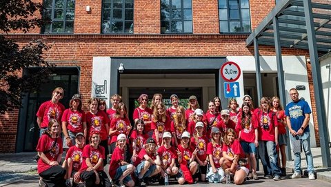 Na zdjęciu grupa młodzieży a w tle budynek Fabryki Aktywności Miejskiej. Po prawej stronie w niebieskiej koszulce Opiekun grupy.
