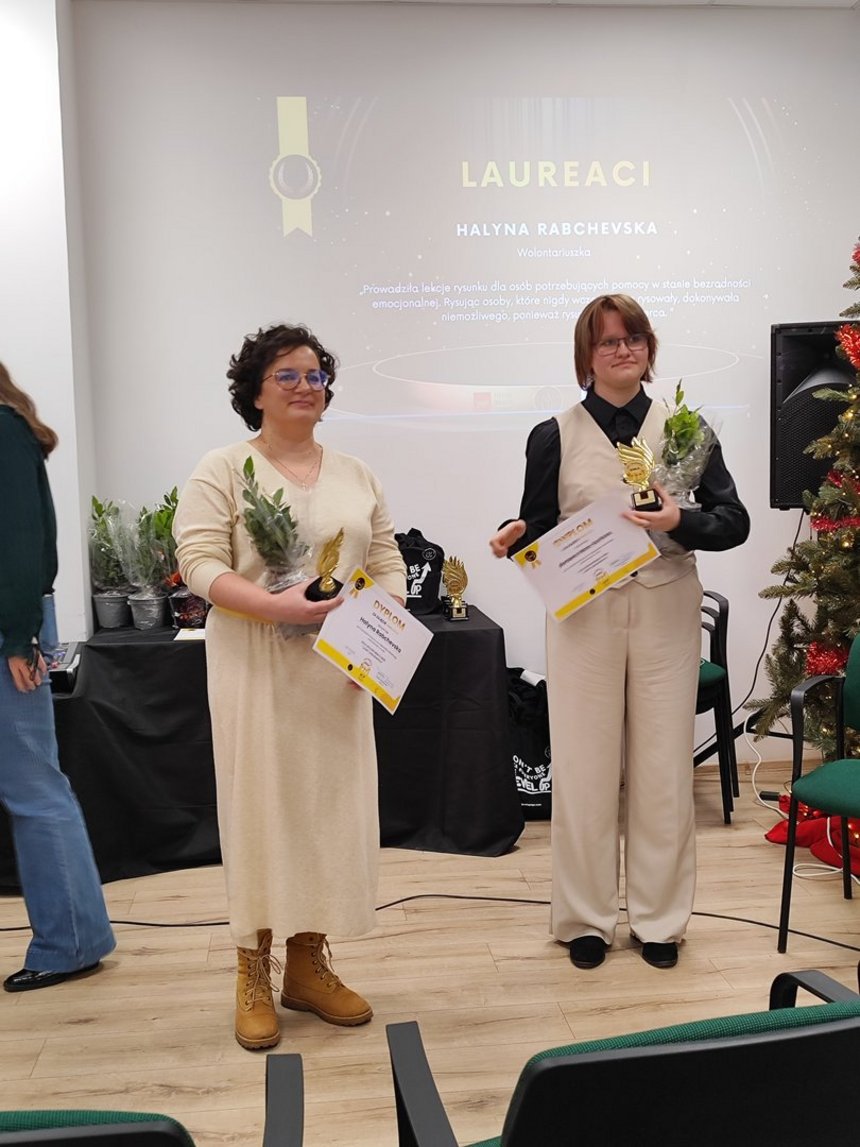 Na zdjęciu widzimy dwie osoby w jasnych strojach trzymajace w dłoniach statuetki, kwiaty oraz dyplomy. Zdjęcie zrobione w pomieszczeniu na tle białego ekranu.