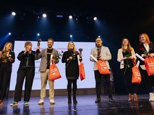 Na zdjęciu widać siedem osób stojących obok siebie na scenie. W tle znajduje się duży biały ekran. Pięć osób trzyma w dłoniach pomarańczowe torby z białym napisem „Moje zakupy niosą pomoc” oraz czerwone róże. Trzy z nich dodatkowo puchar laureata. Uczestnicy są zwróceni przodem do widza i pozują do wspólnego zdjęcia.