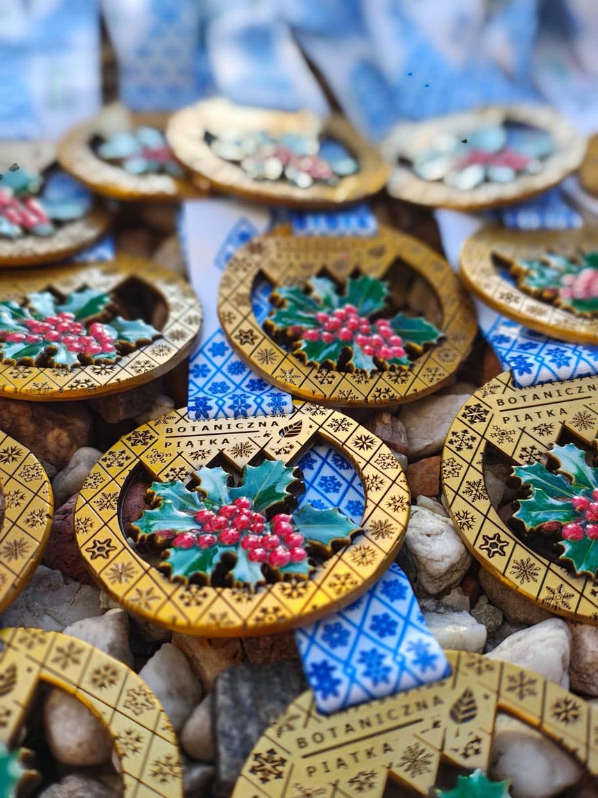 Zdjęcie przedstawia kolorowe medale w złotym kolorze z niebieską tasiemką. W centralnym punkcie medalu znajduje się zielony liść z owocami żurawiny.